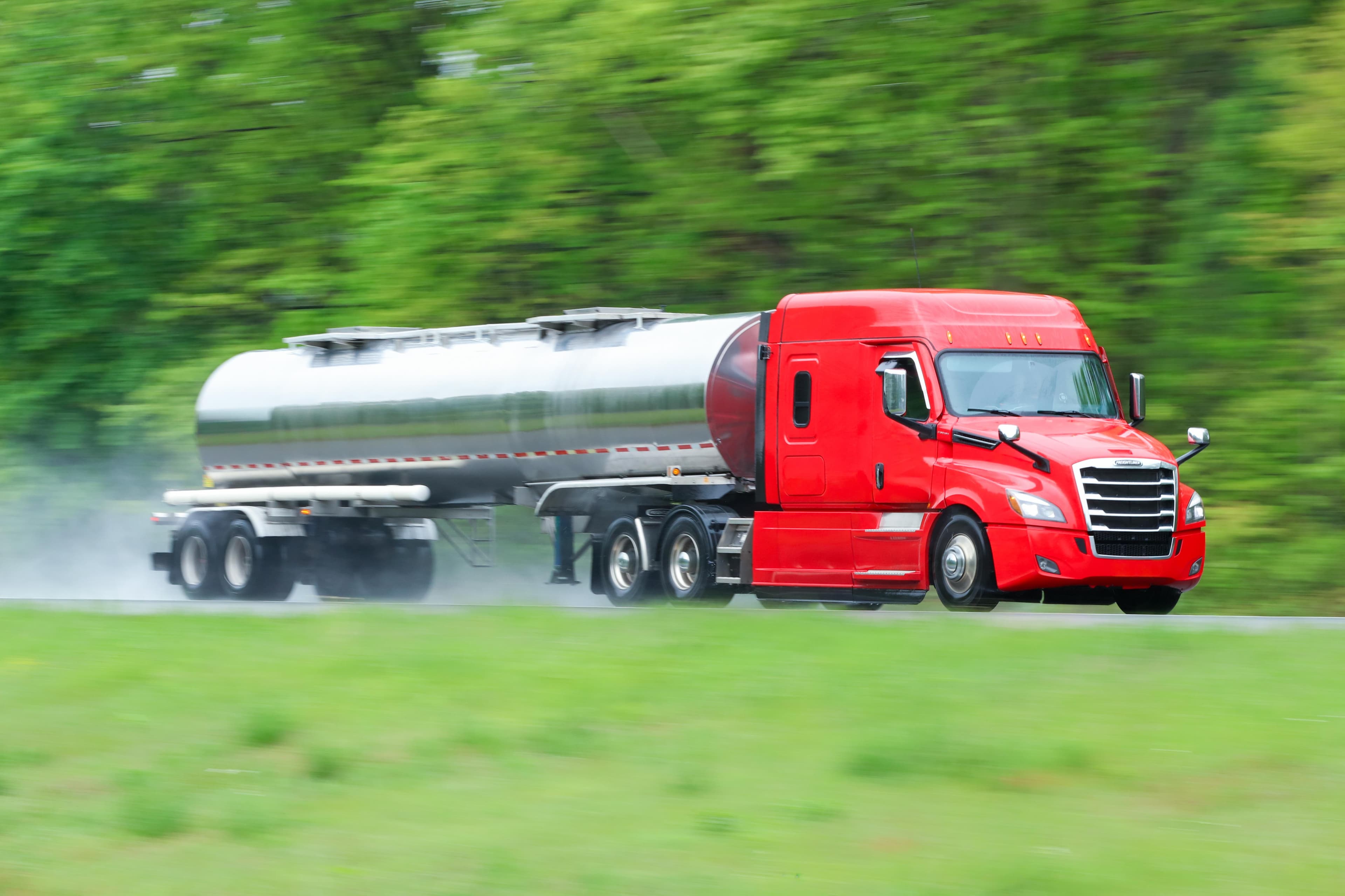 Red semi-truck pulling a silver tanker trailer driving on a highway with motion blur effect showing speed, surrounded by lush green vegetation