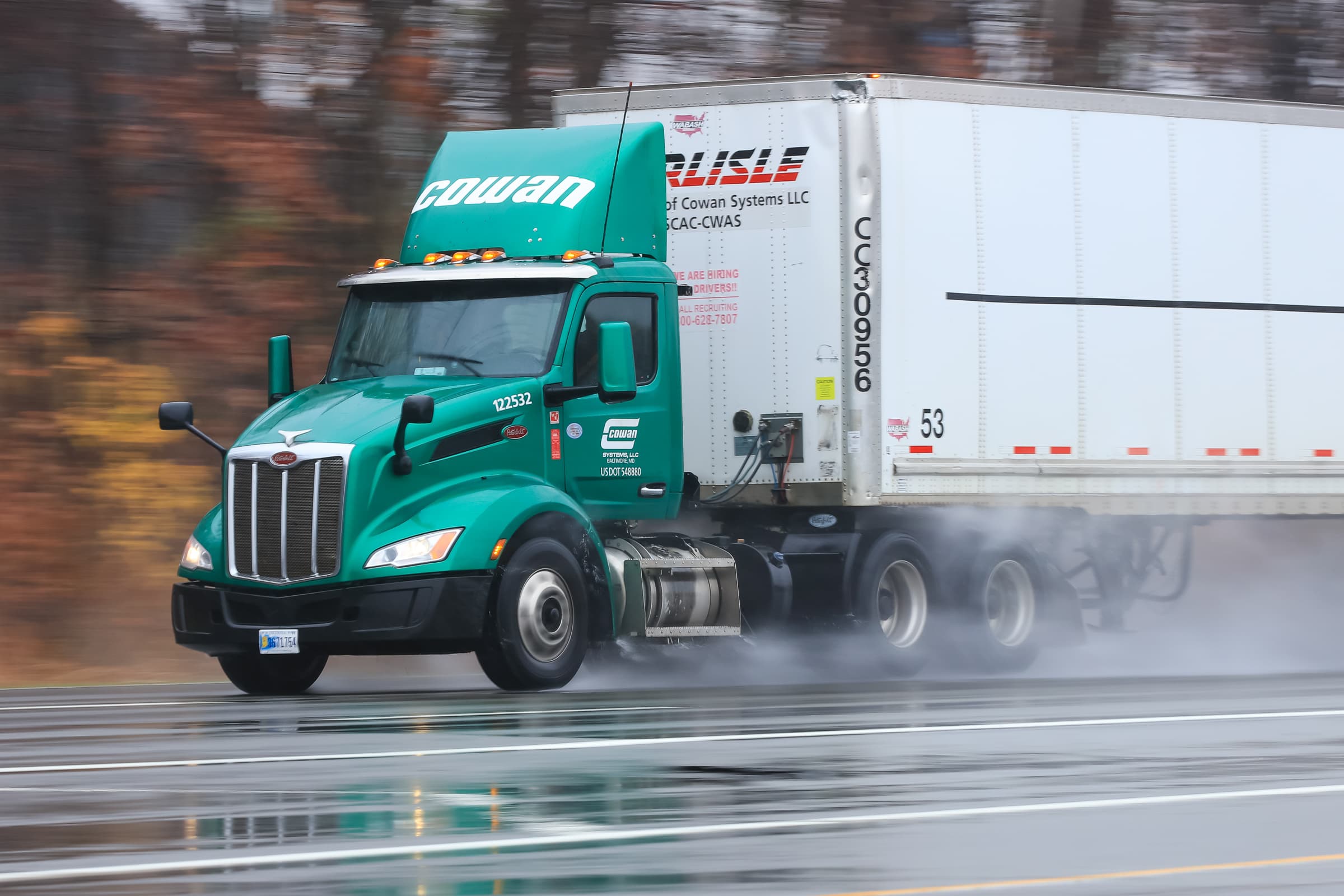 Green Peterbilt truck pulling white Cowan trailer on wet highway with motion blur effect