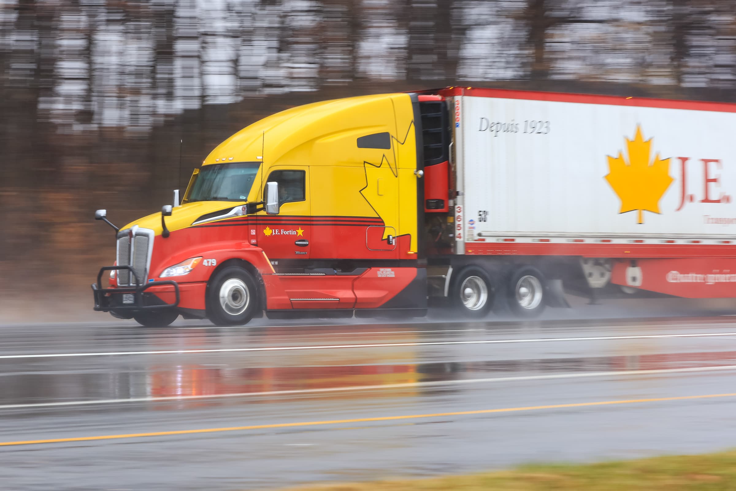 Yellow and red semi-truck with white trailer traveling on wet highway in rainy conditions with motion blur background