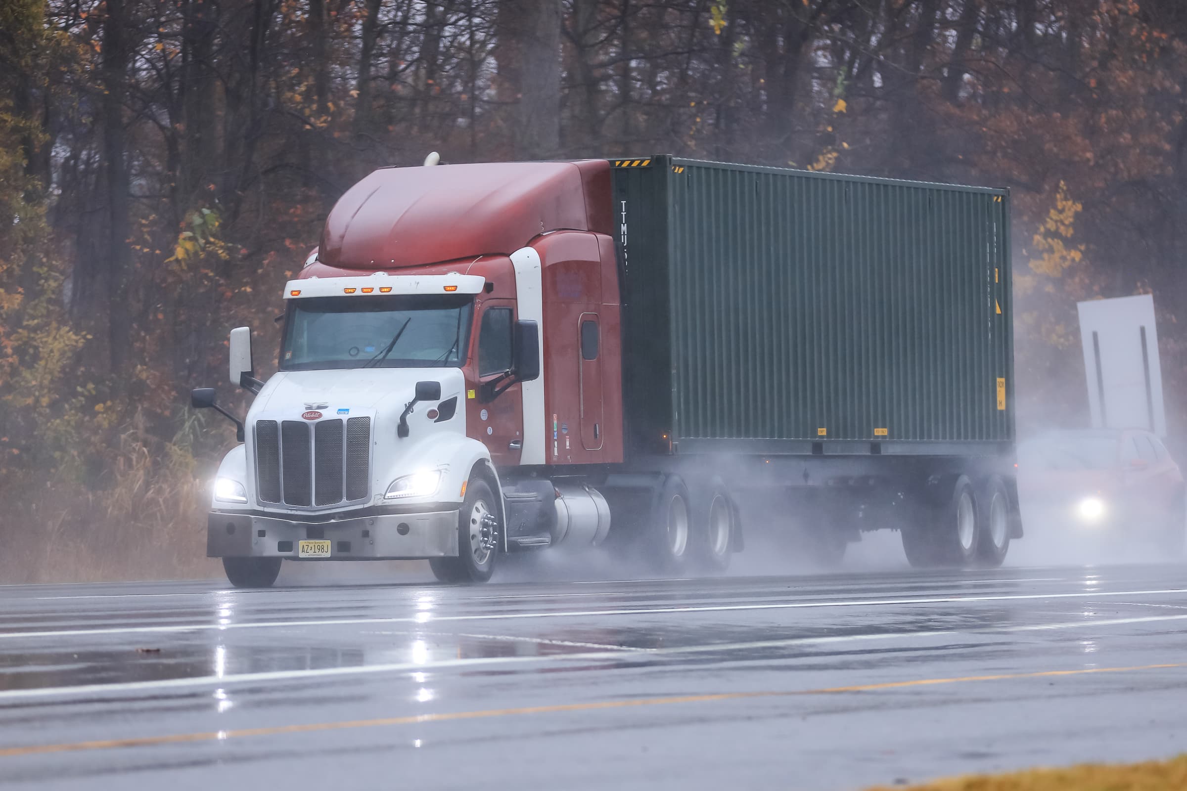 White and red Peterbilt semi-truck pulling a blue shipping container on a wet highway during rainy conditions with autumn trees in the background